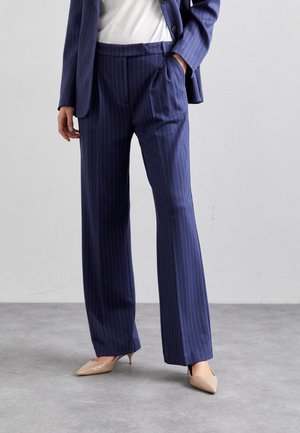 Femme portant un pantalon bleu marine à fines rayures, un blazer assorti, un haut blanc et des talons beiges à bout pointu, debout sur un sol gris.