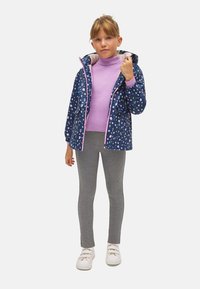 Chaqueta de patrones navy con acentos y forro rosa, llevada sobre un suéter de cuello alto lilas, leggings grises y zapatillas blancas.