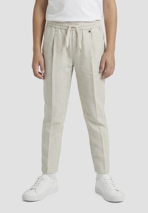 Persona che indossa pantaloni beige con coulisse e pieghe frontali, sneakers bianche e una maglia bianca su uno sfondo chiaro e uniforme.