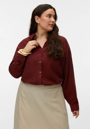 Femme aux longs cheveux noirs portant une chemise bordeaux à boutons, une jupe beige, des bracelets beiges, regardant vers sa droite sur un fond blanc uni.