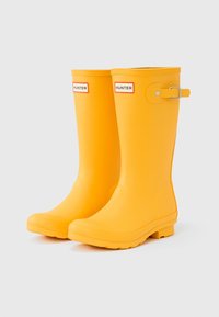 Κίτρινα λαστιχένια μπότες βροχής με στρογγυλεμένη μύτη, υφή στην επιφάνεια, ρυθμιζόμενο λουράκι και αντίθετο λογότυπο σε μαύρο και κόκκινο στο μπροστινό μέρος.