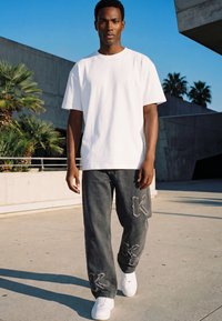 Joven caminando al aire libre con una camiseta blanca, vaqueros oscuros con letras en parches y zapatillas blancas bajo un cielo azul despejado.