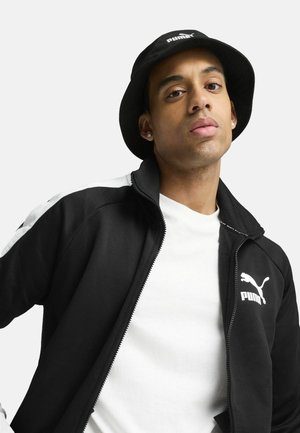 Joven con sombrero tipo bucket negro de Puma, chaqueta negra de Puma con una franja blanca y camisa blanca, mirando confiado a la cámara sobre un fondo liso.