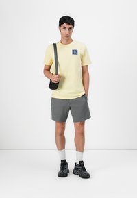 Camiseta de algodón amarilla con un parche de logo azul, pantalones cortos grises con un patrón texturizado, bolso cruzado negro, calcetines blancos, zapatillas deportivas negras.