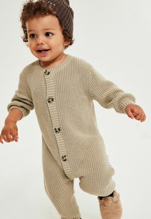 Beige strikket heldragt med rund halsudskæring, lange ærmer, knaplukning foran og ribstrikkede manchetter, kombineret med brun strikket hue.