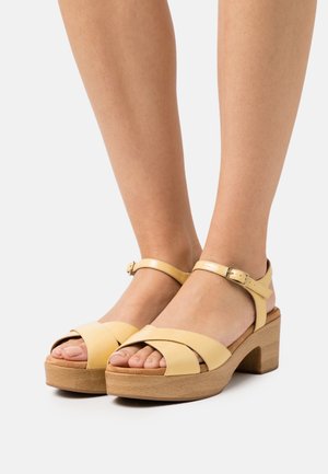 Des pieds portant des sandales plateformes en cuir jaune clair avec des lanières croisées et une boucle à la cheville sur des talons en bois.