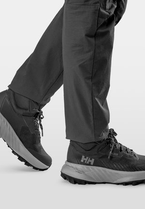 Czarne wodoodporne buty turystyczne z grubą, bieżnikowaną podeszwą, noszone z czarnymi spodniami, pokazane w trakcie kroku na białym tle.