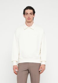 Felpa color crema con collo a polo, polsini a costine e una piccola toppa con logo sul petto. Texture morbida, vestibilità rilassata e design minimalista.