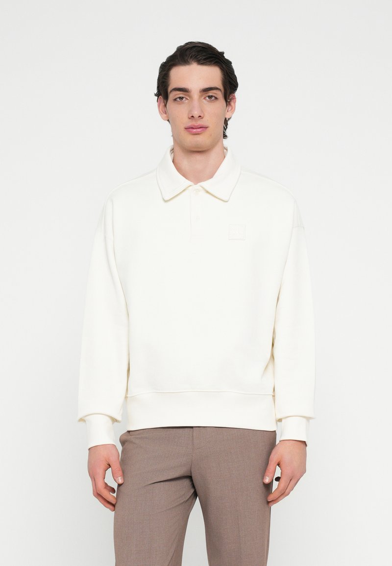 Felpa color crema con collo a polo, polsini a costine e una piccola toppa con logo sul petto. Texture morbida, vestibilità rilassata e design minimalista.