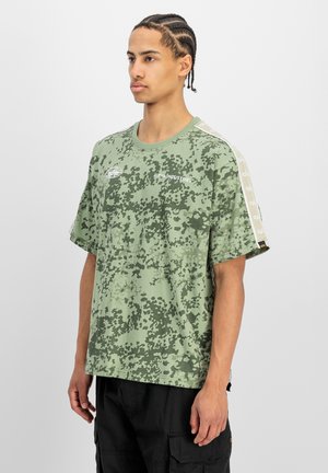Jonge man met gevlochten haar die een groen camouflagepatroon Alpha Industries T-shirt en zwarte cargobroek draagt, staand tegen een effen achtergrond.