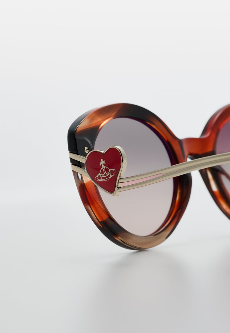 Braune und schwarze oversized Sonnenbrillen mit verlaufenden Gläsern, goldenen Details und einem roten Herzelement an der Bügel, mit grafischem Design.