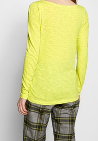 Haut léger à manches longues de couleur jaune néon vif, avec un décolleté large et un tissu texturé, associé à un pantalon à carreaux gris.