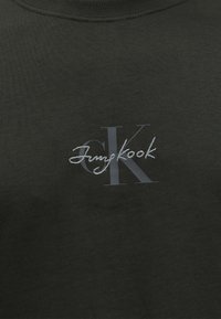 Σκούρο ύφασμα με μεγάλο γκρι λογότυπο "CK" και λευκό κειμενικό κείμενο "Jungkook" κεντημένο πάνω στα γράμματα.
