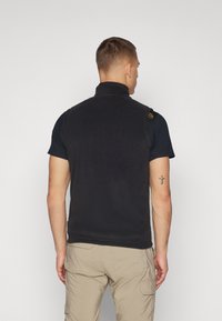 Gilet en polaire noir avec un col montant et des poches latérales, présentant un design simple et un petit logo sur l'épaule gauche.