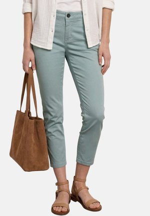 Kobieta ubrana w jasne zielone spodnie do połowy łydki, białą koszulę, beżowe sandały z paskiem wokół kostki i niosąca dużą brązową zamszową torbę.