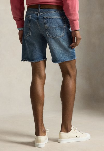 Férfi kopott szegélyű kék farmer rövidnadrágot, fehér sportcipőt és piros hosszú ujjú felsőt visel, semleges háttér előtt áll.