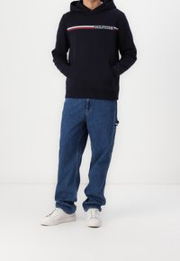Felpa navy con logo e strisce orizzontali, abbinata a pantaloni in denim blu e sneakers bianche, con tasca a marsupio e vestibilità rilassata.