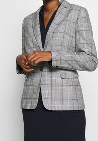 Blazer de cuadros en gris, negro y azul claro. Presenta un diseño estructurado, cuello con solapa, dos bolsillos frontales y un cierre de botón único.