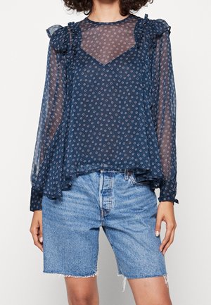 Kobieta ubrana w granatową przezroczystą bluzkę z długimi rękawami, z małymi kwiatowymi wzorami i falbanami, zestawioną z jasnoniebieskimi szortami dżinsowymi.