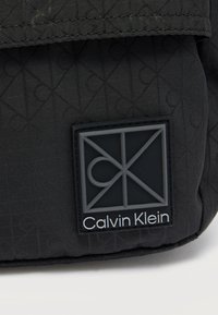 Μαύρη υφασμάτινη τσάντα με ραμμένο σχέδιο, που διαθέτει ένα ανάγλυφο λογότυπο Calvin Klein σε αντίθετο γκρι χρώμα, τονίζοντας γεωμετρικά μοτίβα και κομψό σχεδιασμό.