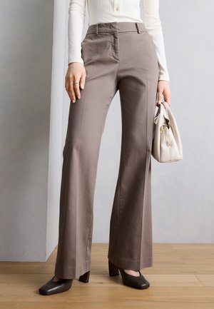 Γυναίκα που φοράει φαρδιά παντελόνια ταφτ, λευκό μακρυμάνικο τοπ, μαύρα τακούνια, κρατώντας μια λευκή τσάντα, στέκεται σε ξύλινο πάτωμα μπροστά από γκρι τοίχο.