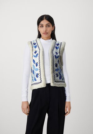 Borduurgilet met een crème kleurige wollen buitenkant, voorzien van blauwe bloemenpatronen en zigzagsteken. Draag het over een witte coltrui, met een losse pasvorm.