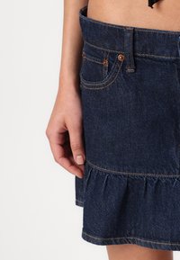 Jupe en denim foncé avec un ourlet à volants évasés, accentuée par des coutures orange contrastantes. Elle présente des poches latérales et une taille ajustée.