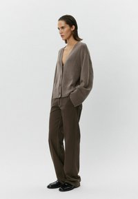 Béžový pletený kardigán s hlbokým V-výstrihom a gombíkmi, skombinovaný s hnedými širokými nohavicami a čiernymi topánkami, stojace na svetlom pozadí.