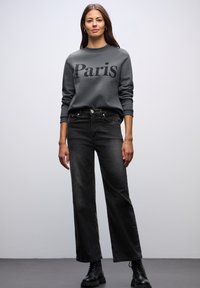 Szary sweatshirt z nadrukiem "Paryż", zestawiony z czarnymi, wysokim stanem dżinsami. Model nosi czarne wiązane botki. Prosty design, swobodny krój.