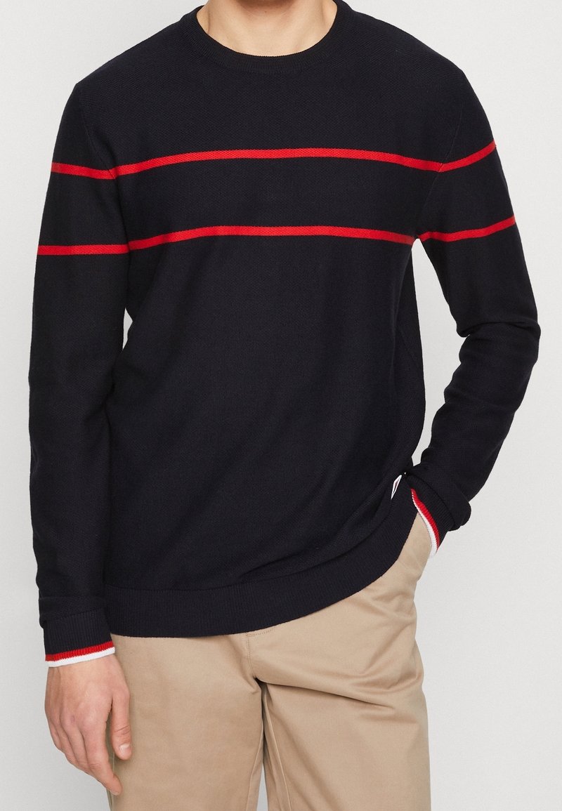Pull en maille marine à rayures horizontales rouges, col rond et poignets côtelés avec accents contrastants. Coupe décontractée portée avec un pantalon beige.