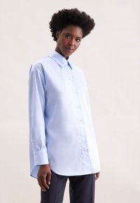 Camicia button-up azzurro chiaro realizzata in tessuto liscio, con colletto classico, maniche lunghe e orlo curvo. Bottoni bianchi discreti.