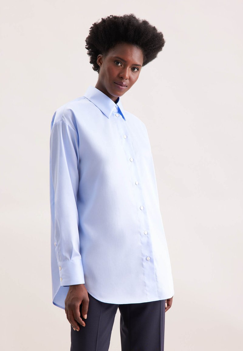 Camicia button-up azzurro chiaro realizzata in tessuto liscio, con colletto classico, maniche lunghe e orlo curvo. Bottoni bianchi discreti.