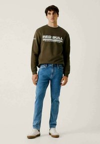 Olive rohelise värvi sviiter, millel on "RED BULL" tekst, koos heledate siniste teksade ja valgete tennisekenadega, millel on pruuni tallaga. Üksik taust.