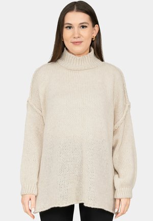 Frau trägt einen locker sitzenden, beigefarbenen, gestrickten Rollkragenpullover mit langen Ärmeln, kombiniert mit schwarzen Hosen, steht vor einem einfarbigen Hintergrund.