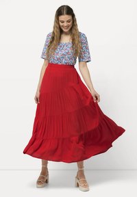 Gonna maxi rossa a strati con un design fluido, abbinata a una maglietta blu a fantasia. Il modello indossa sandali beige con piattaforma. Tessuto leggero, vestibilità comoda.