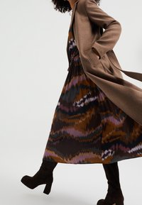 Abrigo de lana marrón sobre un vestido largo estampado con rayas en zigzag en marrón, rosa y negro. La modelo lleva botas negras de ante con tacón grueso.