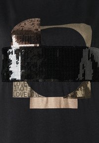 Schwarzes T-Shirt mit abstrakten Formen aus Pailletten und metallischen Akzenten. Enthält ein goldfarbenes metallisches Band und bedruckten Text mit der Aufschrift "LIEBE."