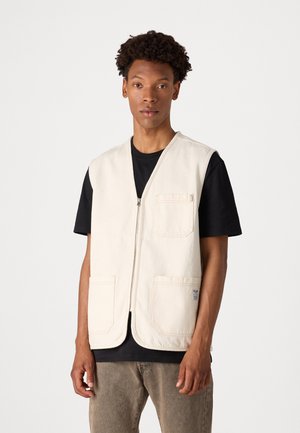 Beige sleeveless vest made of cotton, featuring a front zipper and two large patch pockets. Worn over a black T-shirt.