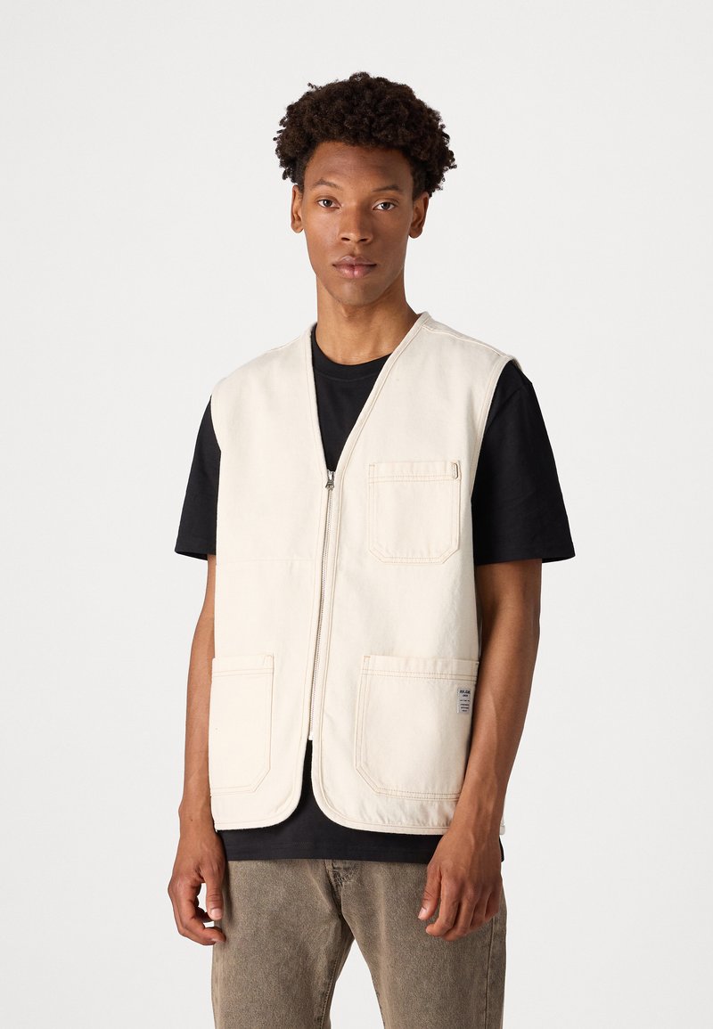 Beige ärmellose Weste aus Baumwolle mit einem Reißverschluss vorne und zwei großen aufgesetzten Taschen. Getragen über einem schwarzen T-Shirt.