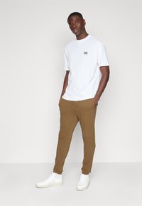 Witte crewneck t-shirt met een klein zwart logo, gecombineerd met bruine joggerpants met zakken en elastische boorden, en witte sneakers.