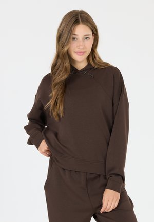 Jonge vrouw met lang haar, gekleed in een bruine hoodie en bijpassende broek, staand met één hand op haar heup tegen een effen achtergrond.