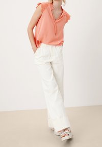 Top corallo senza maniche con pizzo sui lati delle spalle, abbinato a pantaloni bianchi a vita alta con gamba larga e sandali platform grigi chiari.