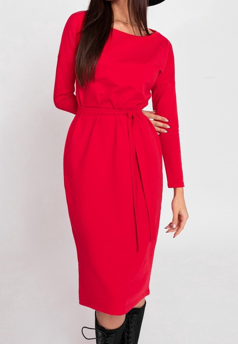 Femme portant une robe midi rouge moulante à manches longues avec une ceinture nouée à la taille et des bottes noires lacées jusqu'aux genoux sur un fond blanc.