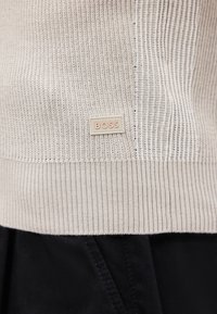 Μπεζ πλεκτή τσάκιση πουλόβερ με ένα μικρό ορθογώνιο λογότυπο BOSS πάνω από μαύρο παντελόνι.