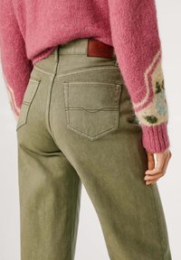 Pantalones de mezclilla verde con un ligero brillo, que cuentan con dos bolsillos traseros y una marca de cuero marrón. Un suéter rosa y esponjoso es parcialmente visible.