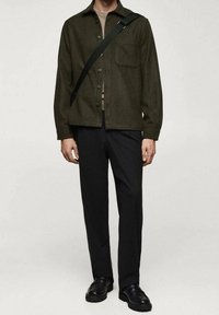 Mørkegrøn uldskjortejakke med krave, forlommer og knaplukning, kombineret med sorte bukser og sorte sko.