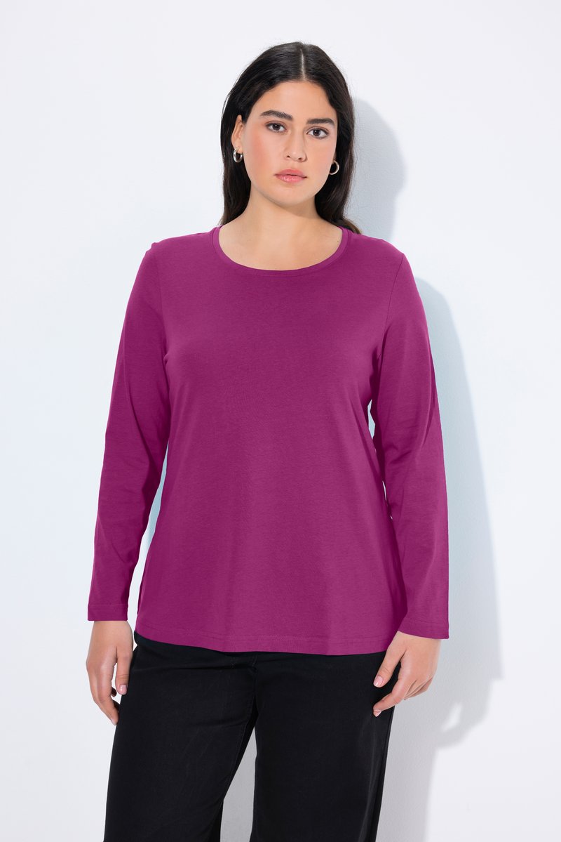 Giovane donna con lunghi capelli scuri che indossa orecchini a cerchio, una maglietta viola a maniche lunghe e pantaloni neri, in piedi su uno sfondo bianco.
