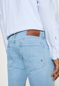 Vaqueros de mezclilla azul claro con ajuste slim, con una pestaña de cuero marrón etiquetada "HILFIGER" y un pequeño logo de color en el bolsillo trasero.