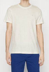 Lys beige kortærmet t-shirt med rund hals, struktureret stof og en lille logo detalje på brystet. Sammen med blå bukser.