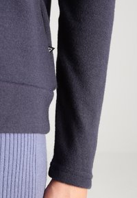 Mörk marinblå långärmad tröja med mudd och sidoseam etikett över ljusblå ribbad stickad byxor mot en ljus bakgrund.